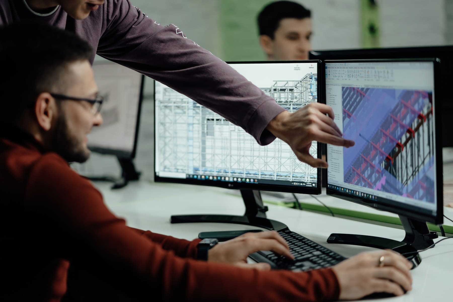 Two engineers at a workstation reviewing a 3D model on dual monitors, one pointing at the assembly on screen while the other works at the keyboard
