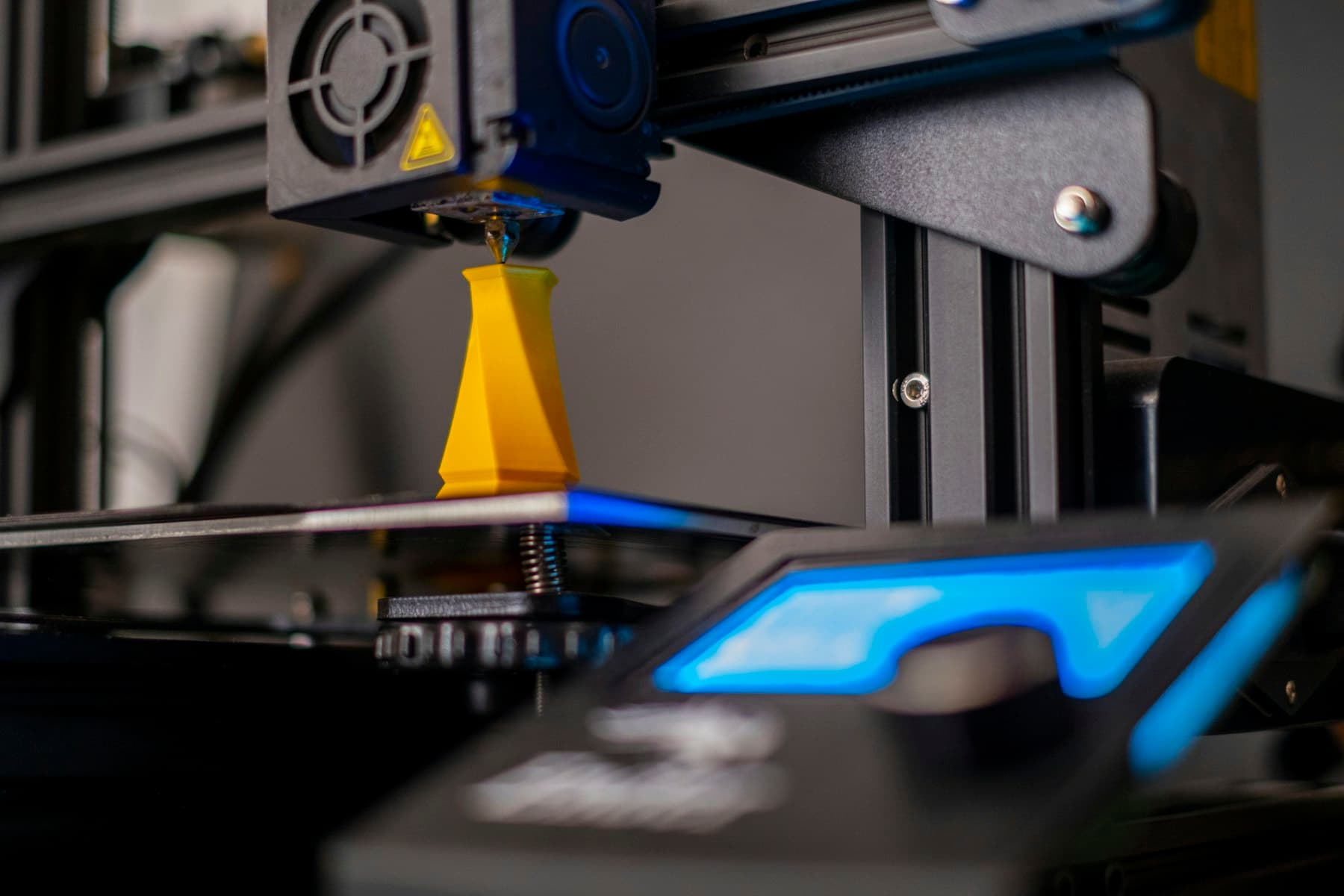 A print head extruding filament onto a part in progress, the machine's control panel lit behind it